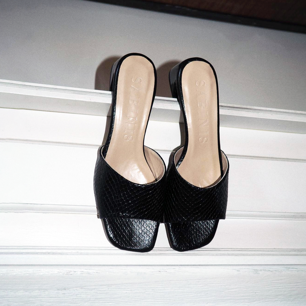 Sandalias mule Essential Black Python de Sabinis, colgadas sobre un panel blanco, con textura de piel negra y plantilla beige, estilo editorial.